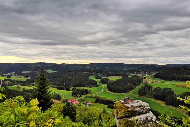 Blick über grüne Hügellandschaft mit Wäldern, verstreuten Höfen und dramatischem, wolkenverhangenem Himmel.