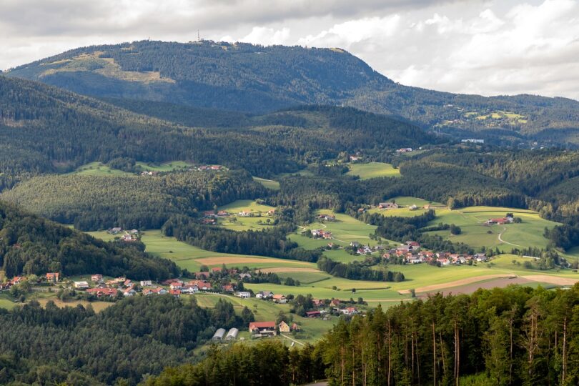 Blick über hügelige Landschaft mit Wäldern, Feldern und verstreuten Dörfern unter teils bewölktem Himmel.