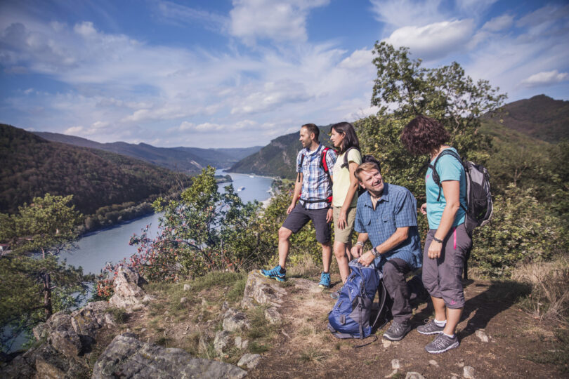Vier Wandernde genießen den Ausblick über ein Flusstal mit bewaldeten Hügeln und blauem Himmel.