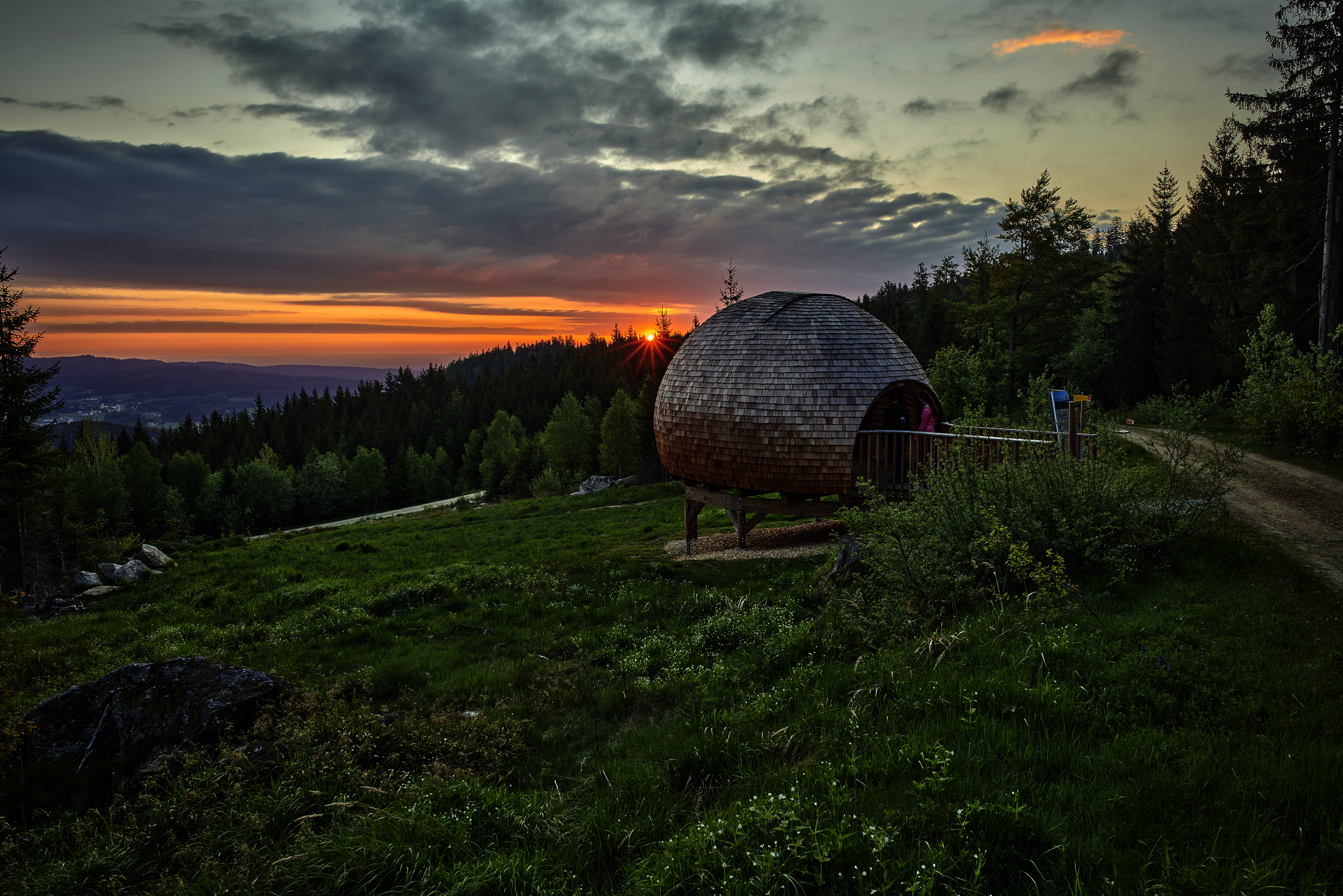 Kugelförmige Holzhütte in Waldlandschaft bei Sonnenuntergang, mit Blick über Hügel und Wälder.