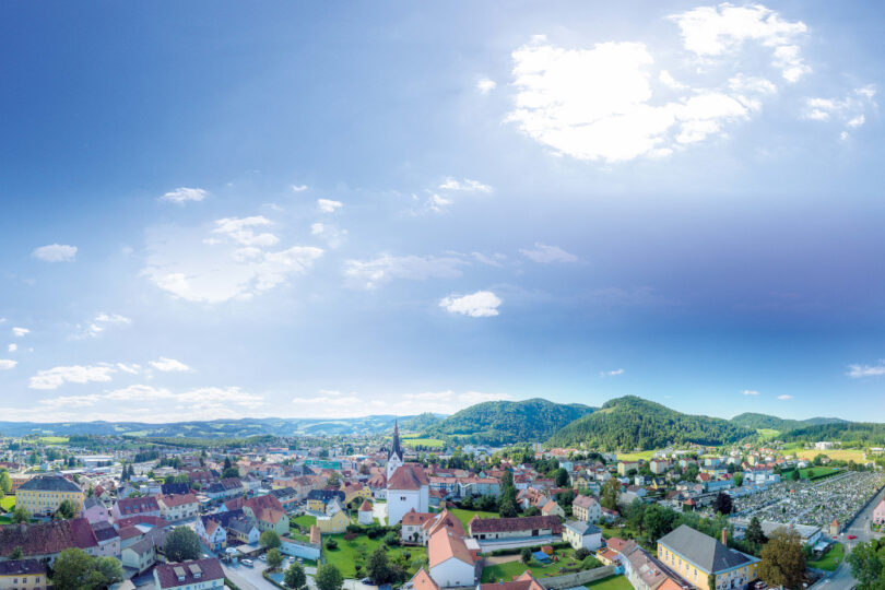 Panoramablick auf eine Kleinstadt mit Kirche im Zentrum, umgeben von Hügeln und teils bewölktem Himmel.