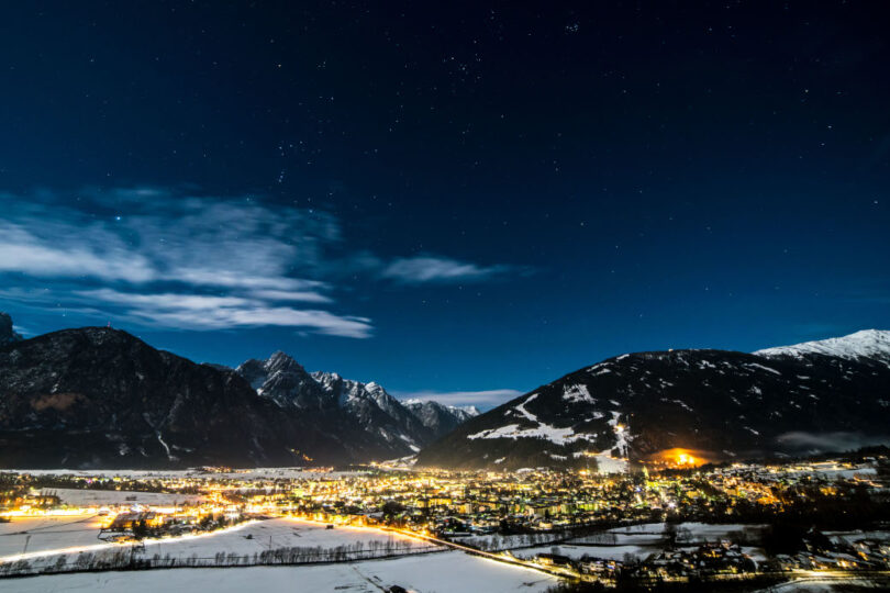 Beleuchtete Winterstadt bei Nacht, umgeben von verschneiten Bergen unter klarem Himmel mit Sternen.