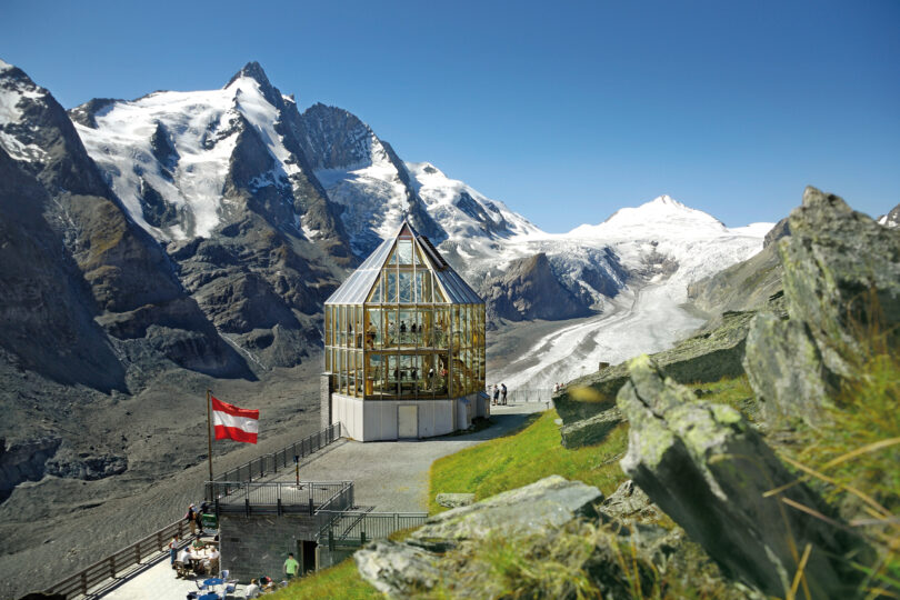 Panoramablick auf Gletscherlandschaft mit moderner Glaspyramide, Großglockner und Österreich-Flagge.