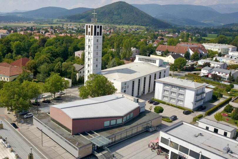 Luftaufnahme eines modernen Kirchenensembles mit hohem Glockenturm, zentralem Kirchenschiff und Vorplatz, umgeben von Wohnhäusern und Hügeln im Hintergrund.