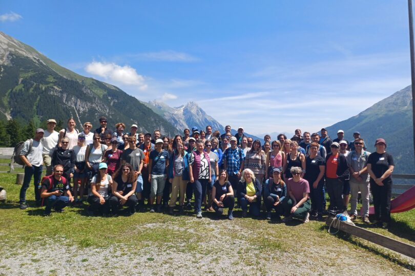Gruppenfoto von KLAR! Manager:innen bei der KLAR! Hauptveranstaltung 2025 in St. Anton, mit etwa 80 Personen im Freien, umgeben von Bergen und Bäumen.