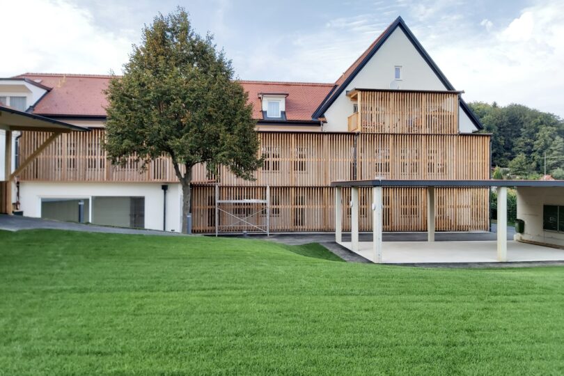 Zweistöckiges Gebäude mit Holzlattenverkleidung, umgeben von Rasenflächen und einem Baum im Vordergrund.