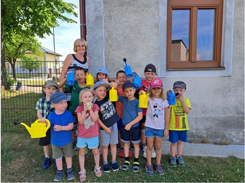 Gruppe von Kindern im Freien, die gelbe Gießkanne halten. Einige tragen Hüte, stehen vor einer Hauswand mit Fenster.