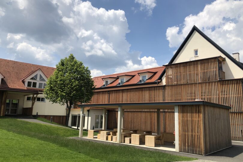 Moderne Gebäude mit Holzverkleidung, darunter ein überdachter Bereich mit Holztischen und -bänken. Im Hintergrund grüne Wiese und Wolken.