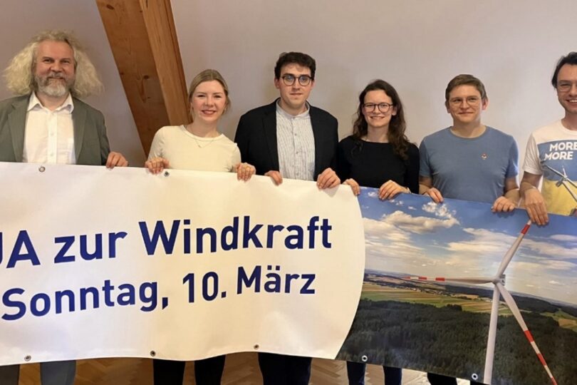 Sechs Personen halten ein Banner mit der Aufschrift 'JA zur Windkraft Sonntag, 10. März'. Im Hintergrund sind Windkraftanlagen und eine Landschaft sichtbar.