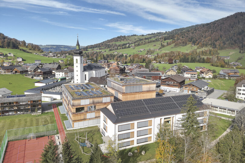 Luftaufnahme eines modernen Schulgebäudes mit Holzfassade und Solarpanelen, umgeben von grünen Hügeln und Wohngebäuden. Im Hintergrund eine Kirche mit einem hohen Turm.