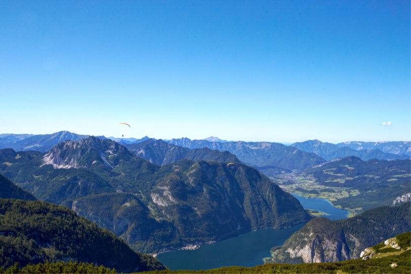 Blick auf eine Berglandschaft mit zwei Gleitschirmen am Himmel, umgeben von Bergen und einem See im Tal.