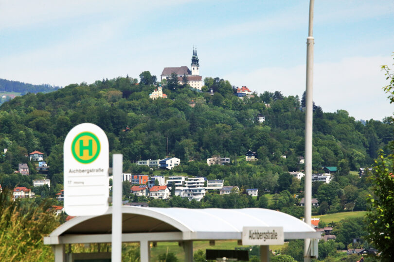 Blick auf einen Hügel mit einer Kirche und einem Turm im Hintergrund, im Vordergrund eine Haltestelle mit dem Schild 'Achterstraße'.