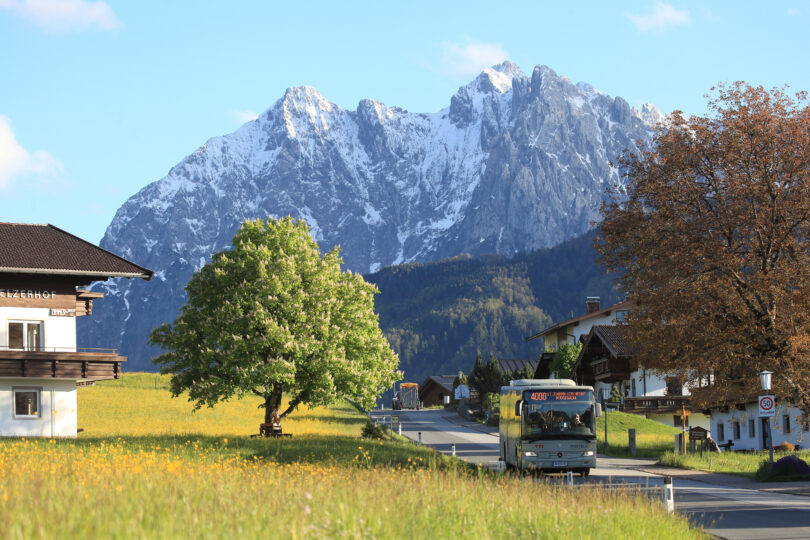 Berglandschaft mit schneebedeckten Gipfeln im Hintergrund, einem Bus der Linie 400, einem Baum und mehreren Gebäuden im Vordergrund.
