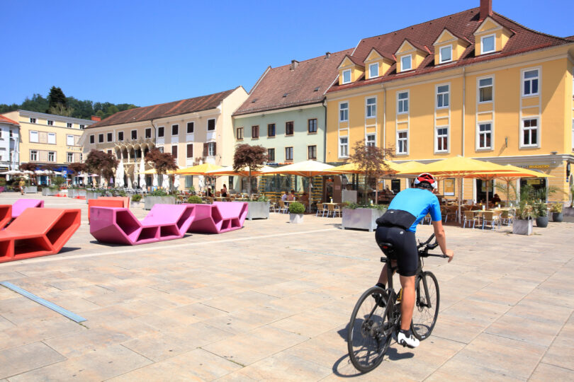 Ein Radfahrer in blauer Kleidung fährt auf einem Platz mit bunten Sitzgelegenheiten in Pink und Rot. Im Hintergrund sind gelbe und weiße Gebäude sowie Sonnenschirme zu sehen.