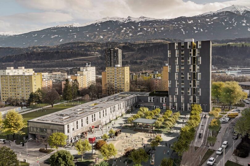 Alois Lugger Platz. Moderne Wohnanlage mit einem hohen, dunklen Turmgebäude und niedrigeren, hellen Bauten, umgeben von grünen Bäumen und einem belebten Platz. Menschen spielen und verweilen in der urbanen Umgebung. Im Hintergrund erstreckt sich eine imposante Berglandschaft mit schneebedeckten Gipfeln, die einen starken Kontrast zur Stadt bildet.