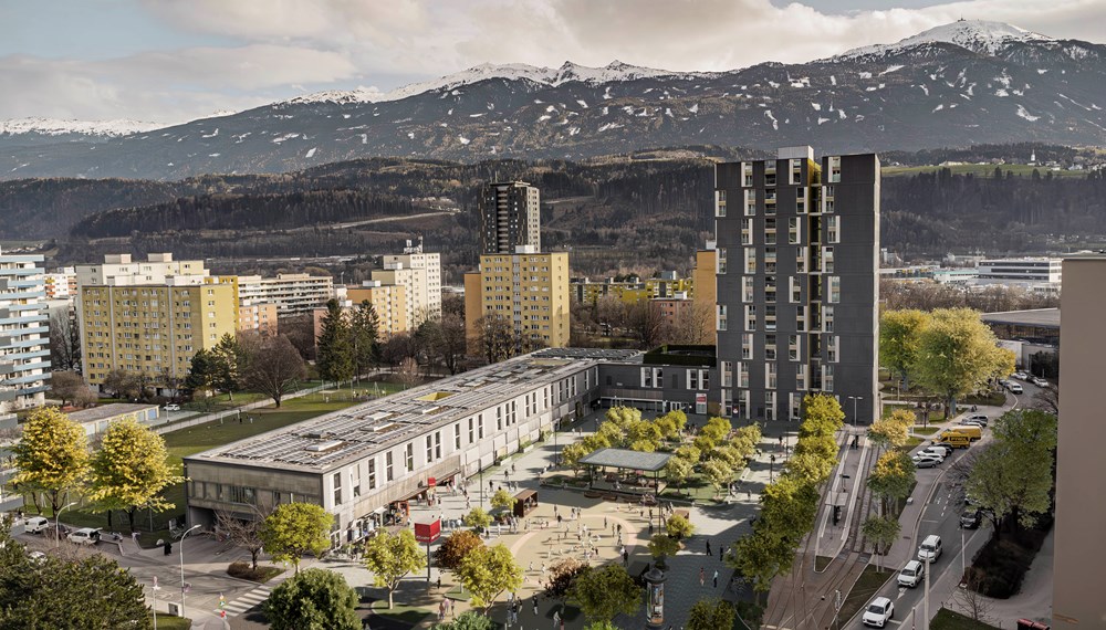 Alois Lugger Platz. Moderne Wohnanlage mit einem hohen, dunklen Turmgebäude und niedrigeren, hellen Bauten, umgeben von grünen Bäumen und einem belebten Platz. Menschen spielen und verweilen in der urbanen Umgebung. Im Hintergrund erstreckt sich eine imposante Berglandschaft mit schneebedeckten Gipfeln, die einen starken Kontrast zur Stadt bildet.