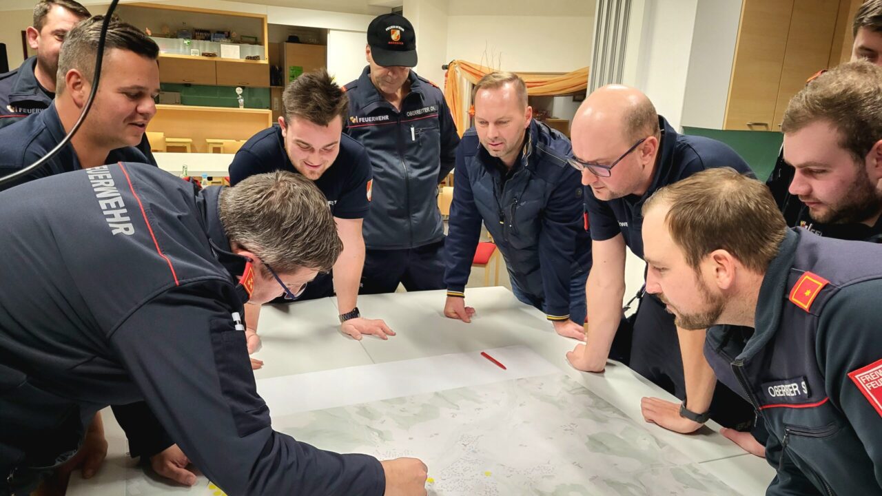 Einsatzvorbereitung Starkregen. Eine Gruppe von Feuerwehrleuten in dunkelblauer Einsatzkleidung steht um einen großen Tisch, auf dem eine detaillierte Karte ausgebreitet ist. Einer der Männer zeigt mit dem Finger auf einen Punkt auf der Karte, während die anderen konzentriert zuhören und die Situation diskutieren. Die Atmosphäre wirkt engagiert und fokussiert, als ob eine Einsatzplanung oder Schulung stattfindet. Der Raum im Hintergrund hat Regale mit Trophäen und eine gemütliche Einrichtung.