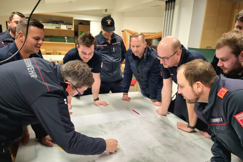 Einsatzvorbereitung Starkregen. Eine Gruppe von Feuerwehrleuten in dunkelblauer Einsatzkleidung steht um einen großen Tisch, auf dem eine detaillierte Karte ausgebreitet ist. Einer der Männer zeigt mit dem Finger auf einen Punkt auf der Karte, während die anderen konzentriert zuhören und die Situation diskutieren. Die Atmosphäre wirkt engagiert und fokussiert, als ob eine Einsatzplanung oder Schulung stattfindet. Der Raum im Hintergrund hat Regale mit Trophäen und eine gemütliche Einrichtung.
