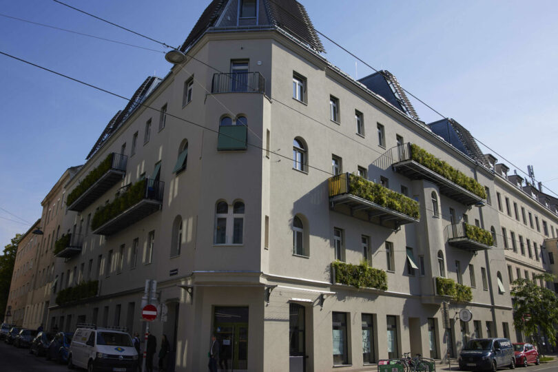 Grauwasseranlage Kauergasse. Modernisiertes, mehrstöckiges Wohngebäude mit nachhaltigen Elementen, darunter begrünte Balkone und große Fensterflächen. Die Dachflächen sind mit Solarpaneelen ausgestattet. Das Gebäude befindet sich an einer Straßenecke in einer urbanen Umgebung mit geparkten Autos, Fahrrädern und Fußgängern.