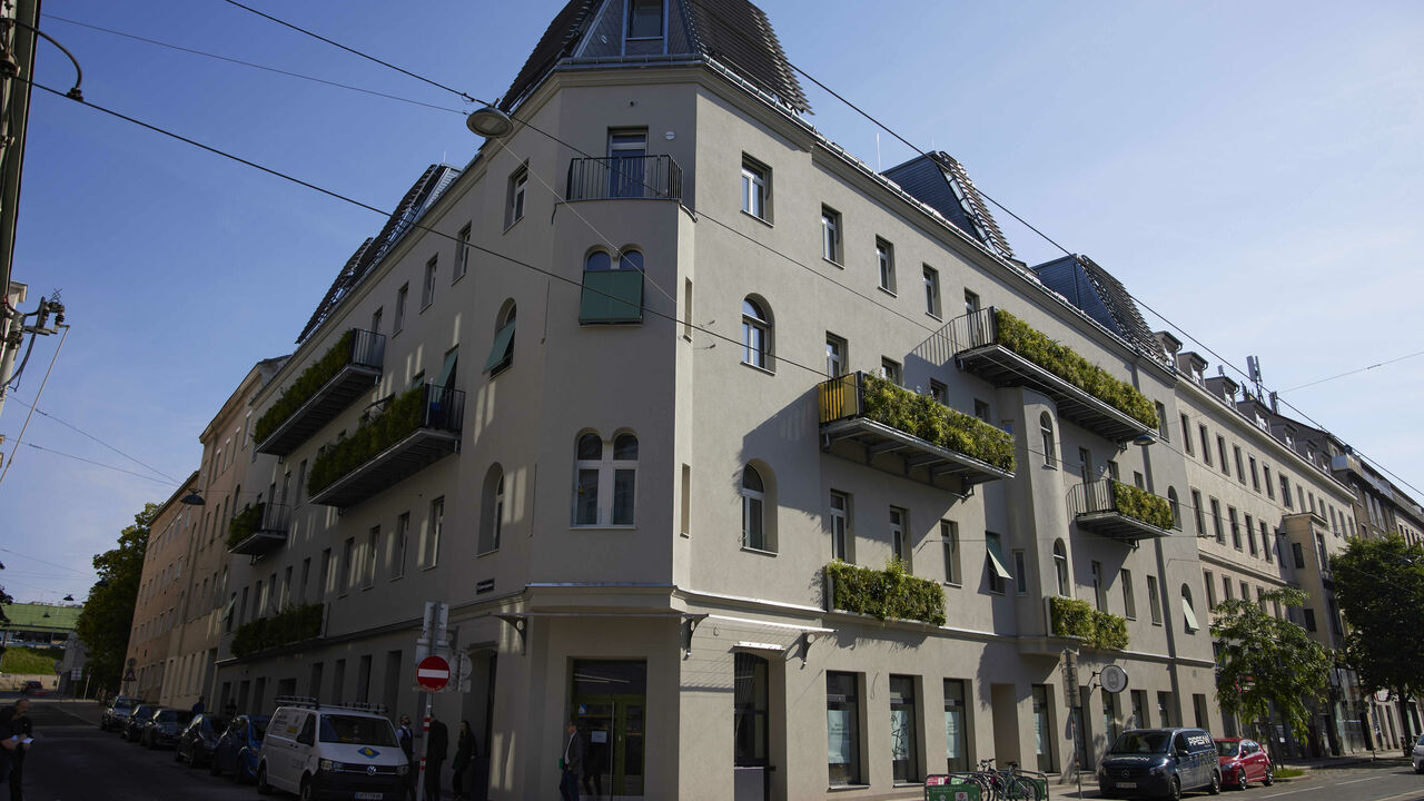 Grauwasseranlage Kauergasse. Modernisiertes, mehrstöckiges Wohngebäude mit nachhaltigen Elementen, darunter begrünte Balkone und große Fensterflächen. Die Dachflächen sind mit Solarpaneelen ausgestattet. Das Gebäude befindet sich an einer Straßenecke in einer urbanen Umgebung mit geparkten Autos, Fahrrädern und Fußgängern.