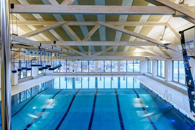 Hallenbad Gänserndorf. Modernes Hallenbad mit einem großen Schwimmbecken mit mehreren Bahnen, umgeben von breiten Gehwegen. An der Decke sind Holzbalken sichtbar, und große Fenster bieten einen Blick auf eine Stadtlandschaft. Links ist ein Kletter- und Balancierparcours über dem Wasser angebracht, rechts eine Wasser-Kletterwand mit bunten Griffen.