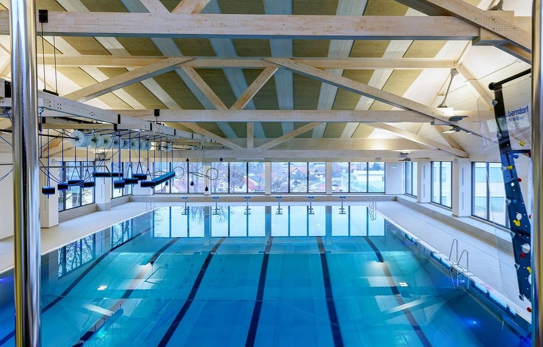 Hallenbad Gänserndorf. Modernes Hallenbad mit einem großen Schwimmbecken mit mehreren Bahnen, umgeben von breiten Gehwegen. An der Decke sind Holzbalken sichtbar, und große Fenster bieten einen Blick auf eine Stadtlandschaft. Links ist ein Kletter- und Balancierparcours über dem Wasser angebracht, rechts eine Wasser-Kletterwand mit bunten Griffen.