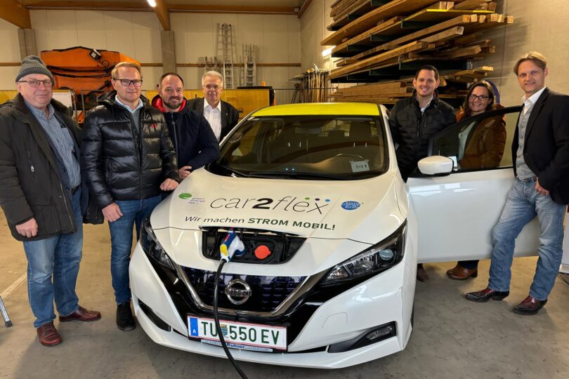 Fünf Personen stehen um ein weißes Elektroauto, das mit 'car2flex' und 'Wir machen STROM MOBIL' beschriftet ist. Das Auto hat eine Ladestation an der Front.