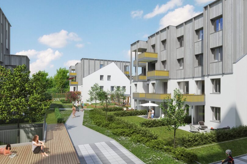 Wohnhausanlage Friedrich-Inhauser-Straße. Moderne Wohnanlage mit mehrstöckigen Gebäuden, die eine grüne Gemeinschaftsfläche umgeben. Balkone mit gelben Akzenten, breite Gehwege und bepflanzte Gärten schaffen eine einladende Atmosphäre. Menschen genießen die Außenbereiche, während Kinder spielen und Fahrrad fahren – eine Szene urbanen Wohnens mit nachhaltigem Flair.