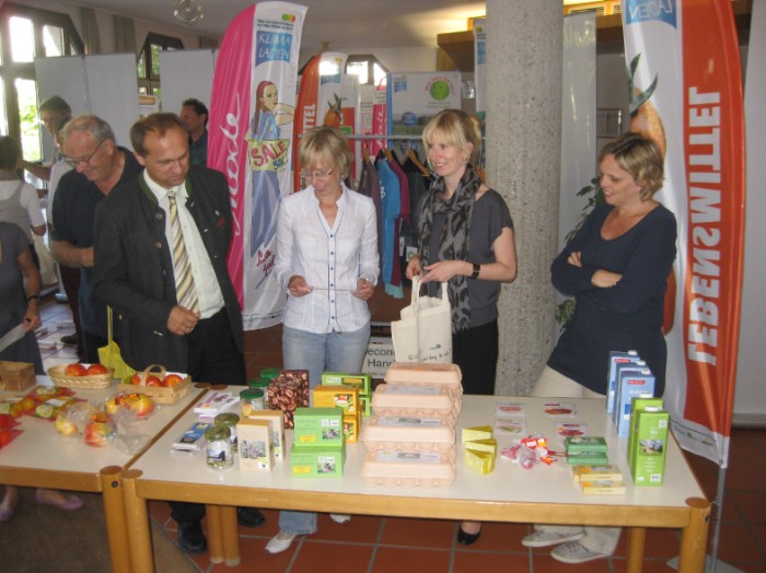 Marktstand mit regionalen Produkten wie Eiern und Äpfeln, dahinter Besucher:innen der Wanderausstellung.