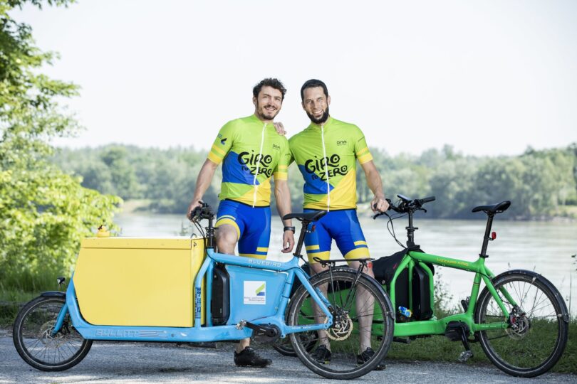 Zwei Männer in Radtrikots posieren mit E-Lastenrädern am Flussufer unter dem Motto Giro to Zero.