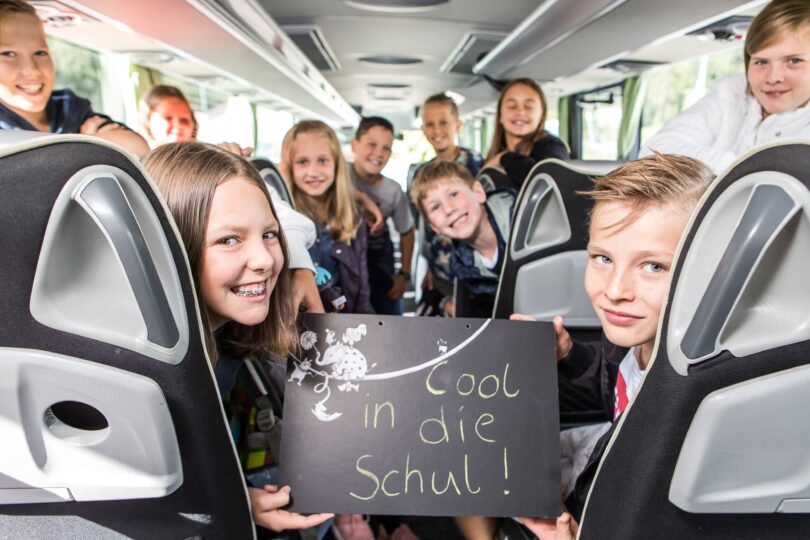 Schulkinder sitzen in einem Bus, ein Mädchen hält ein Schild mit der Aufschrift ‚Cool in die Schul‘.