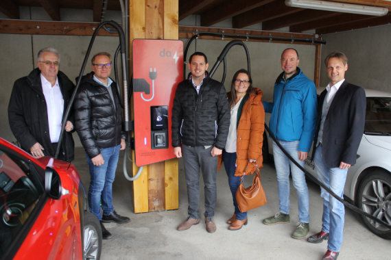 Sechs Personen stehen neben einer roten bidirektionalen Ladestation für Elektroautos in einer Garage.