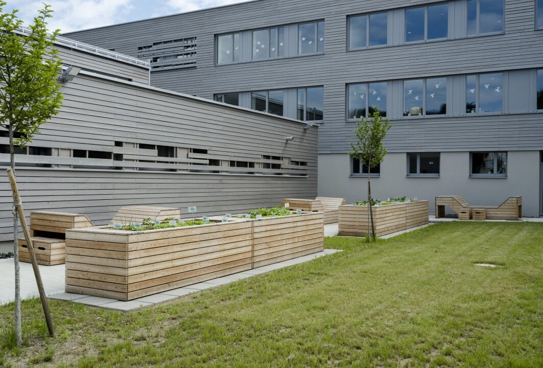 Volksschule Landskron. Moderner, nachhaltiger Innenhof mit Hochbeeten aus Holz, bepflanzt mit jungem Gemüse und Kräutern. Im Hintergrund ein graues Gebäude mit großen Fenstern, teilweise mit aufgeklebten Vogelsilhouetten. Kleine Bäume, eine gepflegte Rasenfläche und Holzliegen laden zum Verweilen ein.