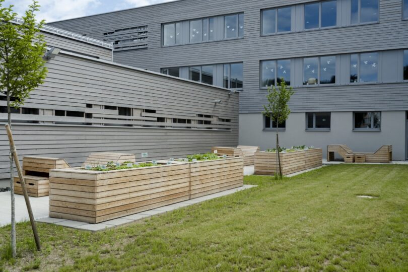 Volksschule Landskron. Moderner, nachhaltiger Innenhof mit Hochbeeten aus Holz, bepflanzt mit jungem Gemüse und Kräutern. Im Hintergrund ein graues Gebäude mit großen Fenstern, teilweise mit aufgeklebten Vogelsilhouetten. Kleine Bäume, eine gepflegte Rasenfläche und Holzliegen laden zum Verweilen ein.