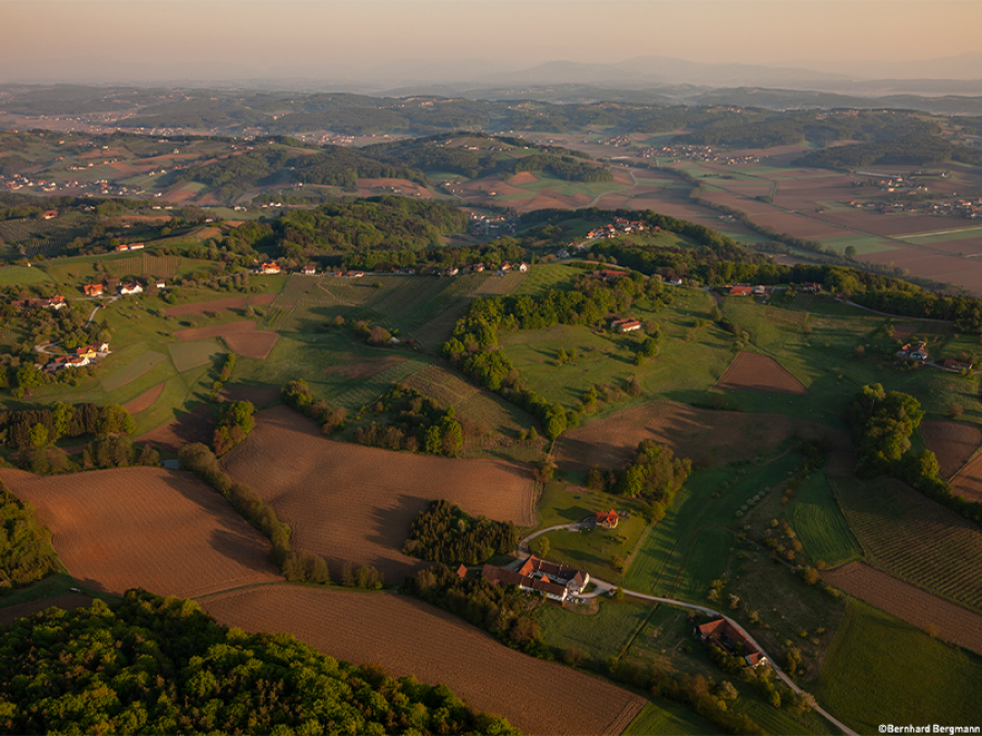 Luftaufnahme einer hügeligen Landschaft mit Feldern und Wäldern. Kleine Dörfer sind verstreut, einige Gebäude sind sichtbar.