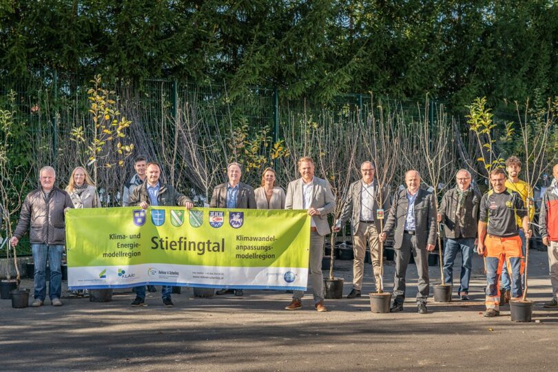 Gruppe von Personen steht vor einer Reihe von Bäumen und Sträuchern. Sie halten ein Banner mit dem Text 'Stiefingtal' und verschiedenen Wappen.