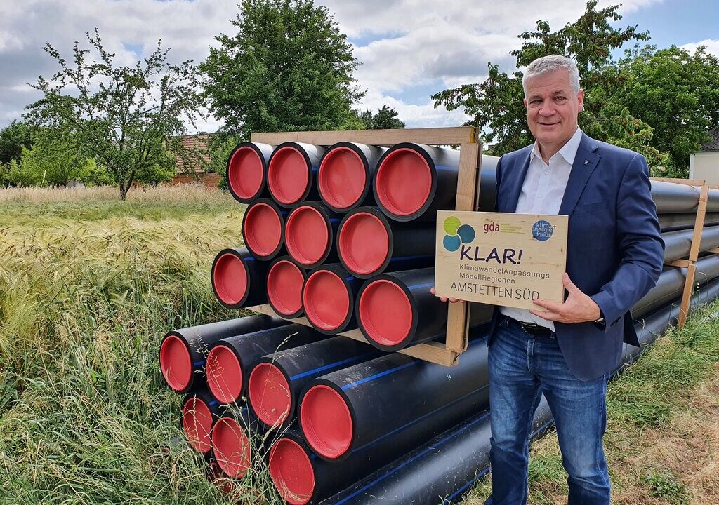 Ein Mann in Anzug steht auf einem Feld neben gestapelten schwarzen Kunststoffrohren mit roten Endkappen und hält ein Schild mit der Aufschrift „KLAR! KlimawandelAnpassungs Modellregionen – Amstetten Süd“. Im Hintergrund sind Bäume, eine Wiese und ein Haus zu sehen.