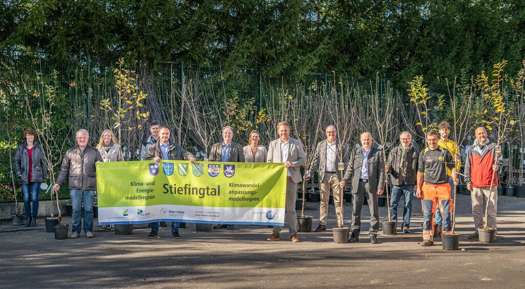 Gruppe von etwa 15 Personen posiert im Freien vor einer Reihe junger Bäume in Töpfen. In der Mitte halten sie ein großes Banner der Klima- und Energiemodellregion Stiefingtal. Die Aktion steht sichtbar im Zeichen klimafreundlicher Begrünung.