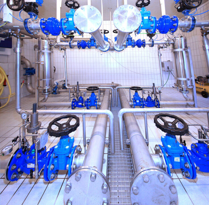 Innenansicht einer modernen Wasseraufbereitungsanlage mit mehreren Edelstahlrohren, blauen Ventilen und Armaturen auf gefliestem Boden. Die Anlage zeigt ein komplexes Rohrleitungssystem mit Absperrschiebern, Druckanzeigern und automatisierten Steuerungen.