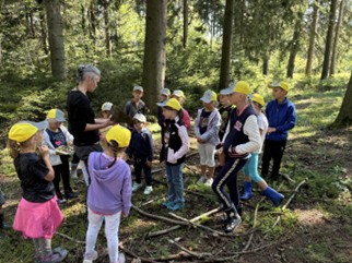Gruppe von Kindern in gelben Hüten, die im Wald stehen. Ein Erwachsener spricht zu ihnen. Bäume und Unterholz im Hintergrund.