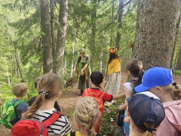 Eine Gruppe von Kindern mit Rucksäcken steht auf einem Waldweg. Zwei Erwachsene in historischen Kostümen, eine mit einer Blumenkrone, interagieren mit den Kindern.