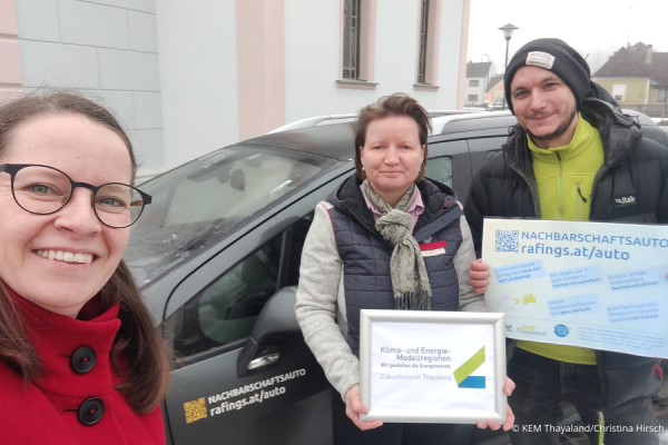 Drei Personen stehen mit KEM-Schildern vor einem Auto.