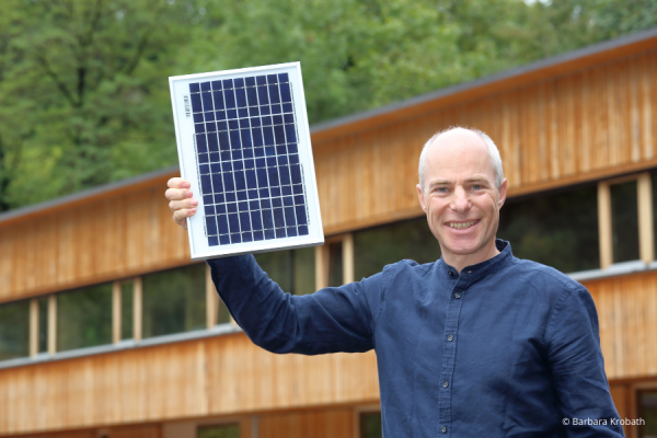 Mann hält PV-Panel in der Hand vor einem Gebäude aus Holz vor Bäumen.