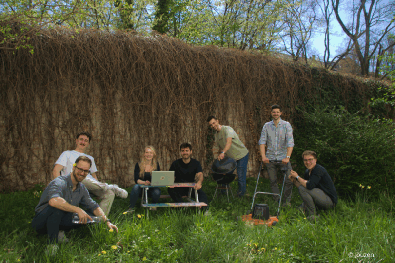 Gruppe an Menschen steht und sitzt in einem Garten vor einer bewachsenen Mauer.