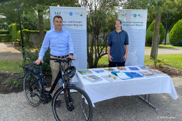 Zwei Personen stehen bei einem Stand mit Rollups draußen vor Bäumen. Der Mann im Bild steht bei einem Fahrrad.