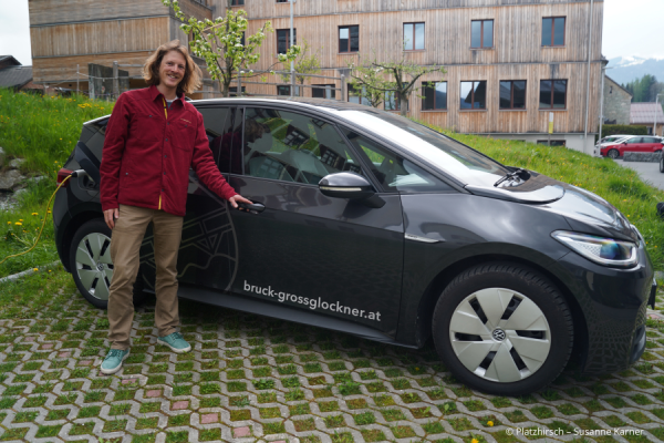 Mann steht vor einem E-Auto auf einem Parkplatz vor einem Holzgebäude.
