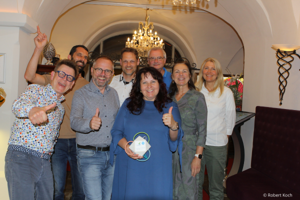 Acht Personen stehen lächelnd in einem eleganten Eingangsbereich mit Kronleuchter.