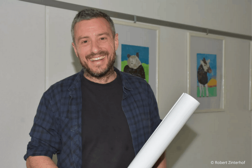 Mann steht mit Papierrolle vor Zeichnungen.