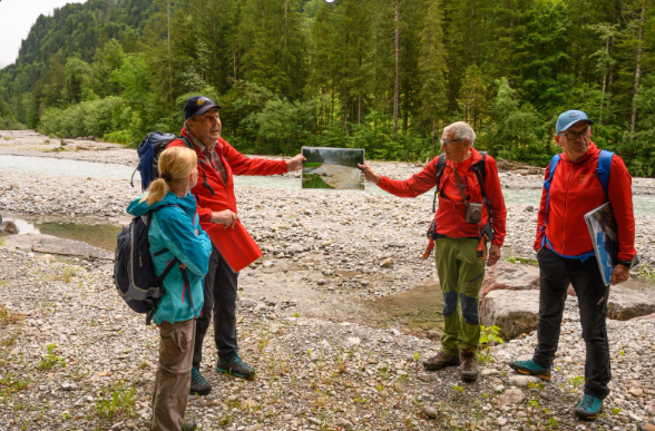 Vier Wanderer in Outdoorbekleidung stehen auf einem Kiesufer, zwei halten eine Karte, im Hintergrund Nadelwald und Flussufer.