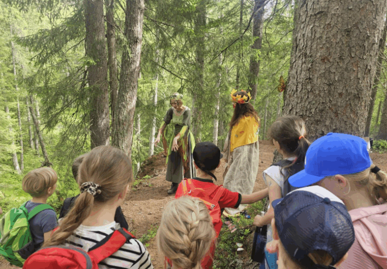Mehrere Kinder mit Rucksäcken stehen im Wald und schauen zwei Personen in Kostümen zu, die auf einem Waldweg stehen.